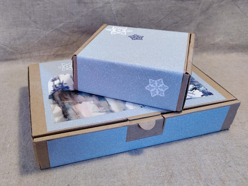 zwei übereinander gestapelte Versandkartons beklebt mit hellblauem Papier und Wintermotiven