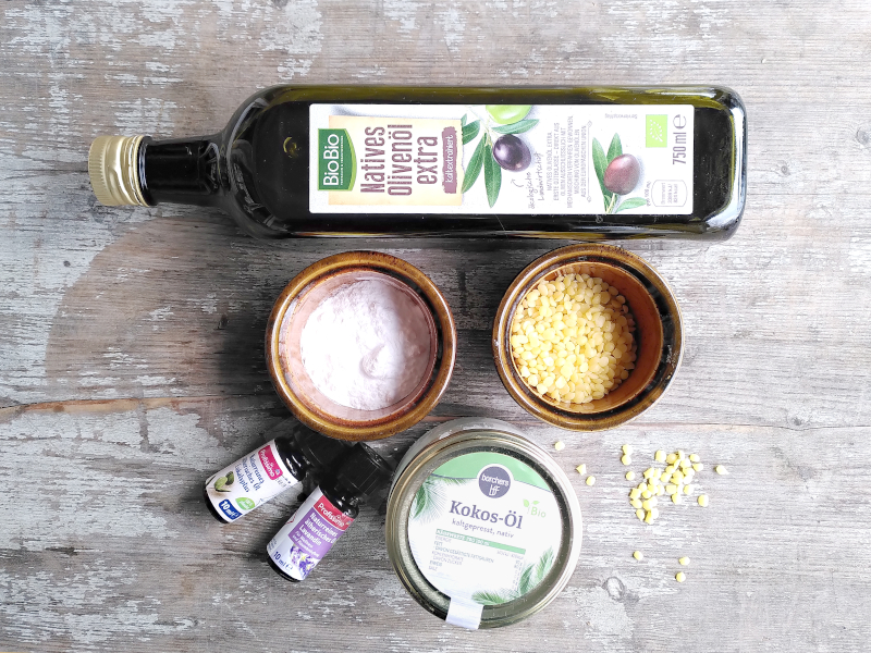 Bio-Olivenölflasche liegt nebn Schüsseln mit Natron und Bienenwachs zwei Flaschen mit ätherischem Öl und einem Glas mit Kokosfett